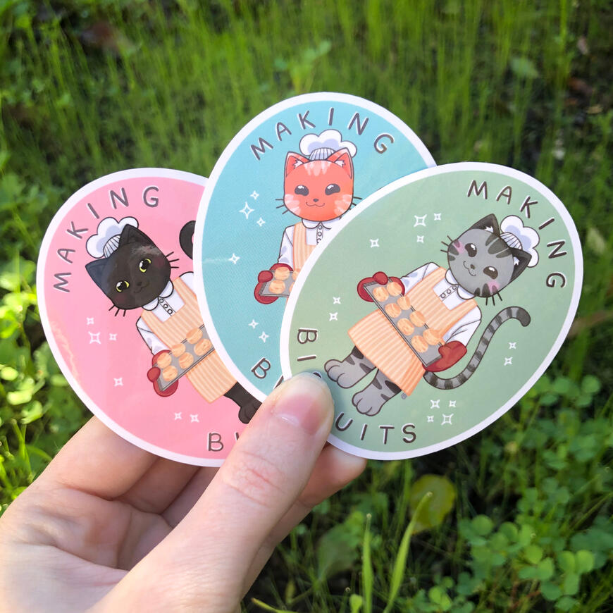 Baking Kitties Image of a hand holding 3 oval shaped stickers, each featuring a different cat wearing a chefs hat, apron, mittens and holding a tray of cookies, the words "Making Biscuits" surrounding the cats. Left to right is: black cat, orange cat, and tabby cat